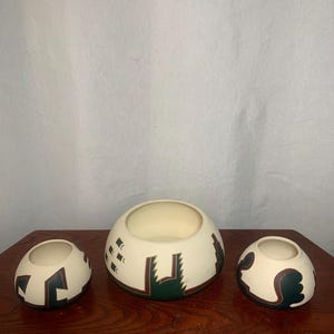 Puede incluir: Tres cuencos de cerámica blanquecina con un diseño del suroeste. Los cuencos tienen forma redondeada y están decorados con patrones geométricos verdes oscuros y marrones. Los cuencos son de diferentes tamaños y están sobre una superficie de madera.