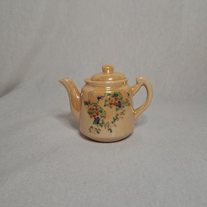 Puede incluir: Una tetera pequeña de color crema con un diseño floral en tonos naranja, verde y azul. La tetera tiene una tapa y un asa.