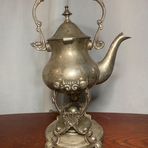 May include: Antique silver-colored tea kettle with ornate detailing. The kettle features a curved spout, a hinged lid, and a decorative handle. It sits on a matching stand with scrolled accents and three feet. The overall design is vintage and elegant.