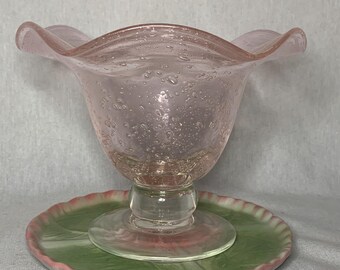 May include: A pink glass bowl with a ruffled rim and a clear glass pedestal base. The bowl has a textured surface with small air bubbles throughout. The bowl is sitting on a green and pink patterned plate.