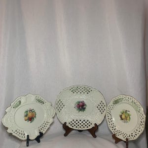 May include: Three white ceramic plates with intricate lace-like designs and fruit illustrations. The plates are on wooden stands.