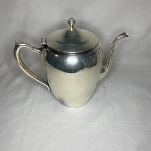 Puede incluir: Una tetera de plata con un acabado brillante y un cuerpo redondeado. La tetera tiene una tapa con un pomo en la parte superior y un pico en el lateral.