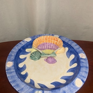 May include: A decorative ceramic plate with a beach theme. The plate features a blue rim with wave designs and a central area with seashells in various colors, including orange, purple, and green. The plate is designed to resemble a sandy beach.