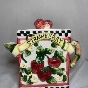 May include: A white ceramic teapot with a strawberry design. The teapot has a square body with a black and white checkered base. The teapot has a handle and spout. The teapot is decorated with strawberries, leaves, and flowers. The word "Strawberry" is written in yellow letters on a yellow ribbon.