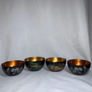 Puede incluir: Cuatro cuencos pequeños negros con bordes dorados y diseños pintados. Los cuencos tienen diferentes diseños, incluyendo flores, pájaros y un dragón.
