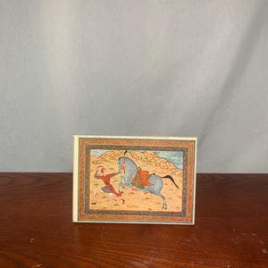 Puede incluir: Una tarjeta rectangular con una pintura en miniatura de una figura con ropa roja persiguiendo a un caballo gris. El caballo tiene una silla de montar roja y corre por un paisaje marrón. La tarjeta tiene un borde decorativo.
