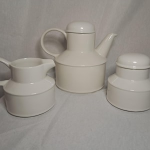 May include: A set of three white ceramic kitchenware items: a teapot, a creamer, and a sugar bowl. The teapot has a lid and a handle. The creamer and sugar bowl have lids.