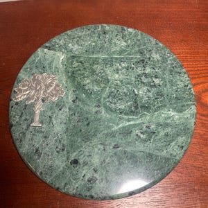 May include: A round, green marble serving board with a silver palm tree emblem. The board has a natural, mottled pattern and a smooth surface. The palm tree is affixed to the board's surface.