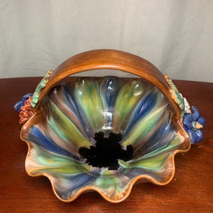 Puede incluir: Una cesta de cerámica con asa marrón y borde festoneado. El interior presenta una mezcla de tonos azules, verdes y amarillos. Los adornos florales adornan los lados, añadiendo un toque decorativo a esta pieza única.