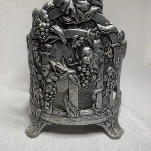 Puede incluir: Candelero de metal plateado con diseño de vid. El candelero tiene tres patas y una base circular.