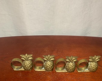 May include: Four brass pineapple napkin rings. Each ring features a detailed pineapple design with a ring for holding a napkin. The napkin rings are arranged in a row on a wooden surface.
