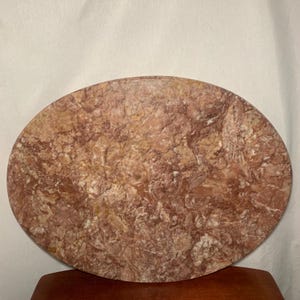 May include: Oval-shaped, reddish-brown marble serving board with a textured surface. The board has a natural stone appearance with variations in color and pattern. The board is likely used for serving food or as a decorative piece.