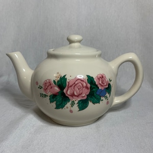 Puede incluir: Tetera de cerámica blanca con un diseño floral con rosas rosas y hojas verdes.