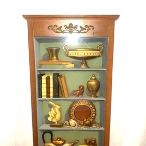 May include: A wooden display shelf with various decorative items. The shelf features gold-coloured vases, books, a plate, a teapot, and a teacup. The background is painted green and blue, with a brown wooden frame.