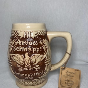 May include: A brown and beige ceramic stein with an eagle and arrows design. The stein says "Arrow Schnapps Schnappsfest 1982" and has a tag that says "Limited Edition Arrow Schnapps Stein".
