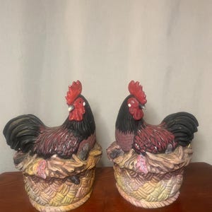 May include: Two ceramic rooster figurines with red combs, black and burgundy feathers, and black tails. The roosters are perched on decorative woven baskets with autumn-colored leaves. The figurines are on a wooden surface.