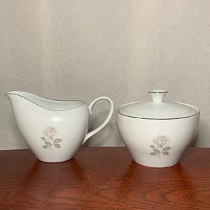 May include: A white ceramic creamer and sugar bowl set. Each piece has a grey rose design and silver trim. The creamer has a handle and spout, while the sugar bowl has a lid with a small knob.