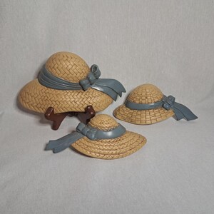 May include: Three straw hats with blue ribbons. The largest hat is on a brown stand. The hats have a woven texture and a light brown color. The ribbons are a muted blue. The hats are decorative.