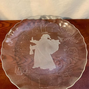 Könnte beinhalten: Ein klarer Glasteller mit einem mattierten Design, das eine Figur in einem langen Mantel und Hut zeigt, die eine Laterne und eine Tasche trägt, vor dem Hintergrund von Häusern und Bäumen. Der Teller hat einen gewellten Rand.