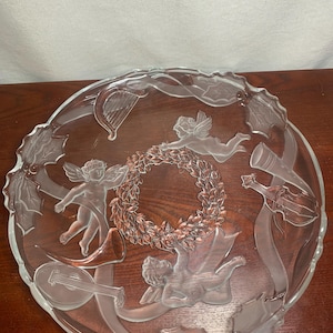 Könnte beinhalten: Transparenter Servierteller aus Glas mit geätztem Design. Er zeigt Cherubim, Musikinstrumente und einen Kranz. Der Teller hat einen gewellten Rand und eignet sich für dekorative oder servierende Zwecke.