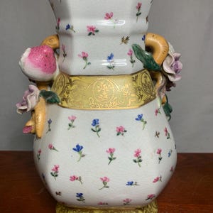 May include: A white porcelain vase with a floral pattern of pink and blue flowers. The vase has a yellow band with gold detailing and is adorned with sculpted flowers and fruit. The vase has a unique shape and is a decorative item.