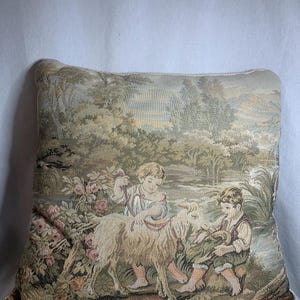 May include: A tapestry pillow featuring a scene of two children playing with a goat in a forest setting. The pillow has a beige background with green, brown, and pink accents.