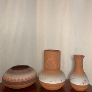 Può includere: Tre vasi in terracotta con disegni geometrici. I vasi variano per forma e dimensione, con una ciotola bassa e arrotondata, un vaso bulboso con collo cilindrico e un vaso sottile. Ognuno presenta accenti bianchi e marroni.