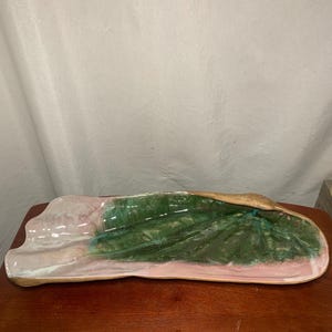 May include: A long, ceramic serving dish with a leaf-like design. The dish features a pink and green colour scheme with a gold-coloured rim. The leaf design is in shades of green, and the dish is on a wooden surface.