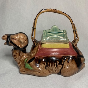 May include: A ceramic camel teapot with a brown, green, and yellow glaze. The teapot has a bamboo handle and a lid with a green glaze.