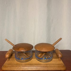 May include: A wooden serving set featuring two glass jars with wooden lids and spoons, resting on a wooden tray. The jars have a blue and yellow floral design. The set is made of wood and glass.