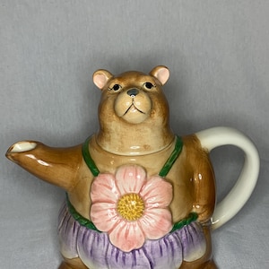 Könnte beinhalten: Eine braune Keramik-Teekanne in Form eines Bären, der einen lila Rock und eine rosa Blume trägt. Die Teekanne hat einen weißen Henkel und einen Ausguss.