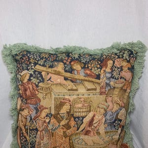 May include: A tapestry pillow with a green fringe border. The pillow features a medieval scene with people in a garden setting. The scene includes a large wooden tub, a wooden structure, and several people in colorful clothing.
