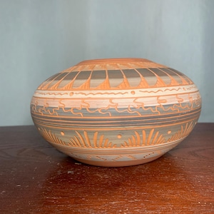 Puede incluir: Un cuenco de cerámica pintado a mano con forma redondeada. El cuenco presenta un diseño de patrones y líneas geométricas de color naranja, gris y blanco. El cuenco está sobre una superficie de madera.