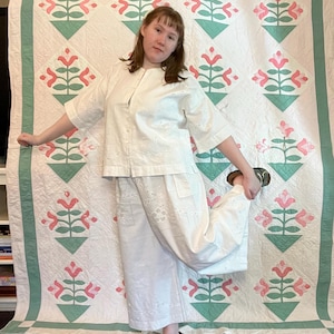 Könnte beinhalten: Ein weißes Ensemble aus Oberteil und Hose mit Lochstickerei. Das Outfit wird mit grünen Sandalen kombiniert. Im Hintergrund befindet sich eine Vintage-Steppdecke mit einem Blumenmuster in Rosa und Grün.