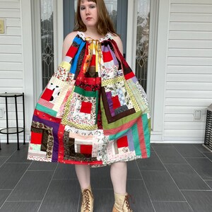 Könnte beinhalten: Ein buntes Patchwork-Kleid mit verschiedenen Stoffmustern und Farben, darunter Rot, Blau, Grün und Weiß. Das Kleid hat einen Neckholder-Ausschnitt und reicht bis zu den Knien. Die Person trägt beige Schnürstiefel.