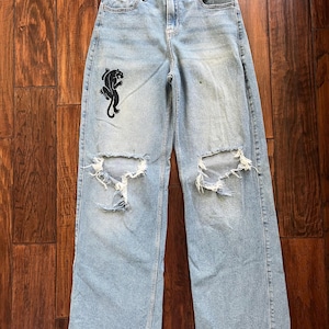 May include: Light blue denim jeans with a high-rise waist, a button and zipper fly, and wide legs. The jeans feature distressed, ripped knees and a black graphic print of a panther on the upper right thigh. The hem is raw and unfinished.