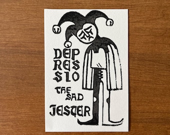 May include: A black and white print of a jester with the text "Depressio The Sad Jester". The jester has a hat with bells and a sad face. The print is on a white rectangular paper.