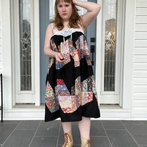 Peut inclure: Robe patchwork avec une jupe noire et un corsage en crochet blanc. La jupe présente des motifs floraux colorés. La robe est associée à des bottines lacées beiges. La tenue est adaptée aux tenues décontractées.