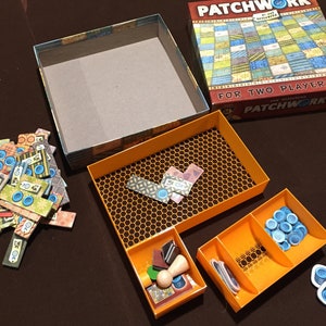 May include: A game box with a patchwork quilt design on the lid. The box is open and the contents are visible. The contents include a game board, game pieces, and a rule book. The game is called "Patchwork 2".