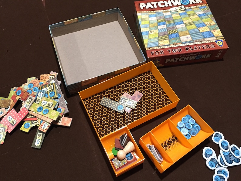 May include: A game box with a patchwork quilt design on the lid. The box is open and the contents are visible. The contents include a game board, game pieces, and a rule book. The game is called "Patchwork 2".