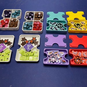 May include: A set of colorful game pieces and trays for a board game. The trays are white with different colored inserts. The game pieces are small and colorful, with different shapes and designs. The game pieces include stars, gears, and animals.