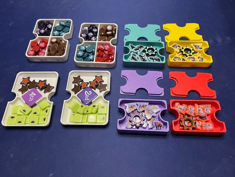 May include: A set of colorful game pieces and trays for a board game. The trays are white with different colored inserts. The game pieces are small and colorful, with different shapes and designs. The game pieces include stars, gears, and animals.