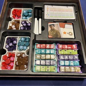 May include: A game board organizer with compartments for storing game pieces, dice, and cards. The organizer is made of black plastic and has a wooden frame. The game pieces are red, brown, purple, and blue. The dice are red, green, blue, and purple. The cards are white with a picture of a man in a military uniform.
