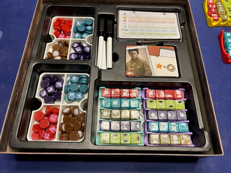 May include: A game board organizer with compartments for storing game pieces, dice, and cards. The organizer is made of black plastic and has a wooden frame. The game pieces are red, brown, purple, and blue. The dice are red, green, blue, and purple. The cards are white with a picture of a man in a military uniform.