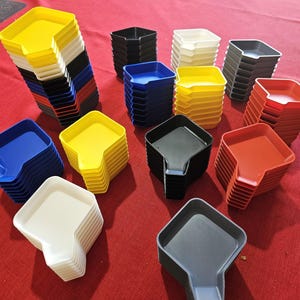 Op de afbeelding: Een verzameling van 11 stapels kleine, vierkante, plastic bakjes met een tuit. De bakjes zijn in verschillende kleuren, waaronder geel, blauw, rood, wit en zwart.