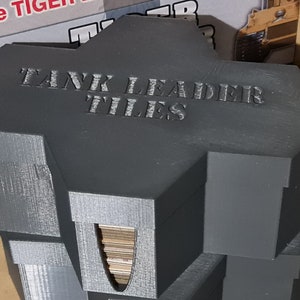 May include: A grey, 3D printed hexagonal box with the text "TANK LEADER TILES" on the top. The box is designed to hold game tiles and has a slot on the side for easy access.