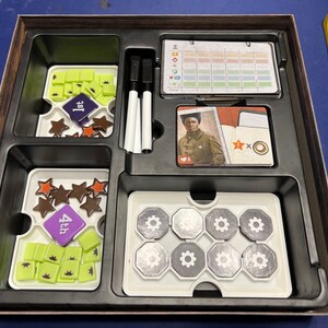 May include: A wooden box with compartments for storing game pieces. The box contains a variety of game pieces, including stars, gears, and small squares. The box also includes a score sheet, a player card, and two white dry-erase markers.