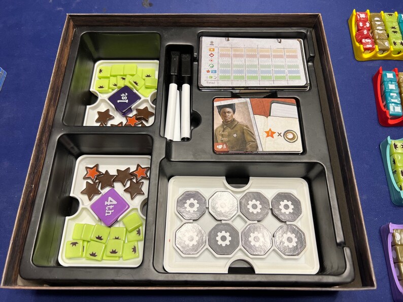May include: A wooden box with compartments for storing game pieces. The box contains a variety of game pieces, including stars, gears, and small squares. The box also includes a score sheet, a player card, and two white dry-erase markers.