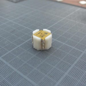 Op de afbeelding: Een klein, wit, kubusvormig object met een gouden bovenkant. De bovenkant heeft een patroon van kleine vierkantjes en de tekst "1234567890".
