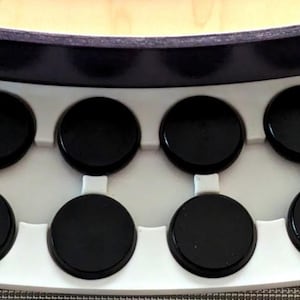 Könnte beinhalten: Ein weißes Tablett mit acht schwarzen, kreisförmigen Pads. Das Tablett ist rechteckig mit abgerundeten Kanten und befindet sich unter einem dunklen, gebogenen Objekt. Die Pads sind gleichmäßig verteilt und scheinen aus glänzendem Material zu bestehen.